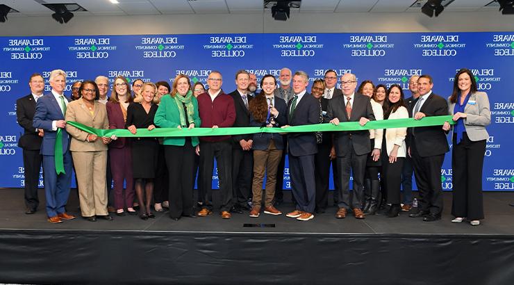 A large group of people standing in front of a blue backdrop with the Delaware Tech logo tiled across the backdrop. There is a long green ribbon spanning the group and Delaware Tech student Maximiliano Rios is holding a pair of large scissors in preparation to cut the ribbon